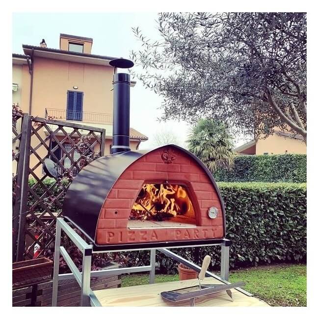 Outdoor Pizzone pizza party wood fired pizza oven on a wheeled metal stand with a black chimney, shown lit with visible flames in a garden patio setting.