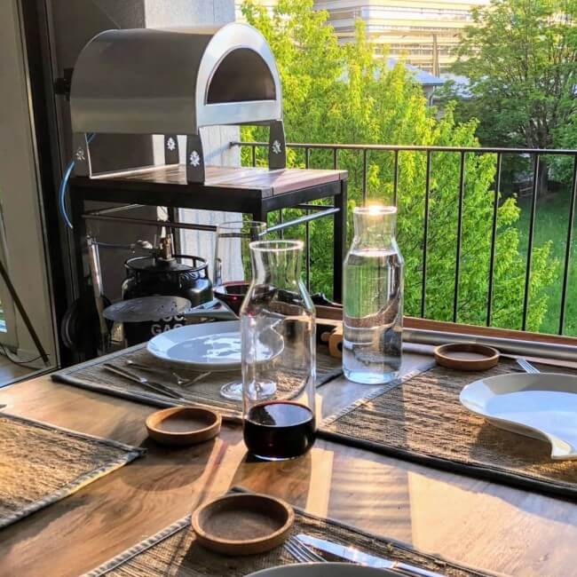 A wooden dining table set with plates, bowls, glass carafes, and a pizza party ardore gas pizza oven on a cooking deck with a balcony railing and green outdoor foliage visible in the background.