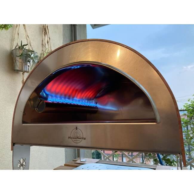 Pizza Party Ardore gas outdoor pizza oven with a arched metal door, shown lit with blue and red flames inside. The PizzaParty logo is visible on the lower front.