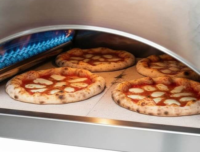 A close-up of four pizzas cooking inside the copper Pizza Party Ispirazione outdoor gas pizza oven.