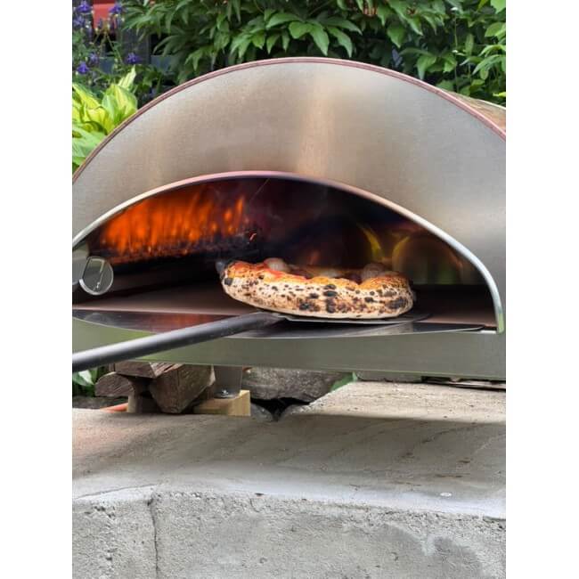 An antique copper Pizza Party Ispirazione outdoor gas pizza oven baking a single pizza in a lush garden setting.