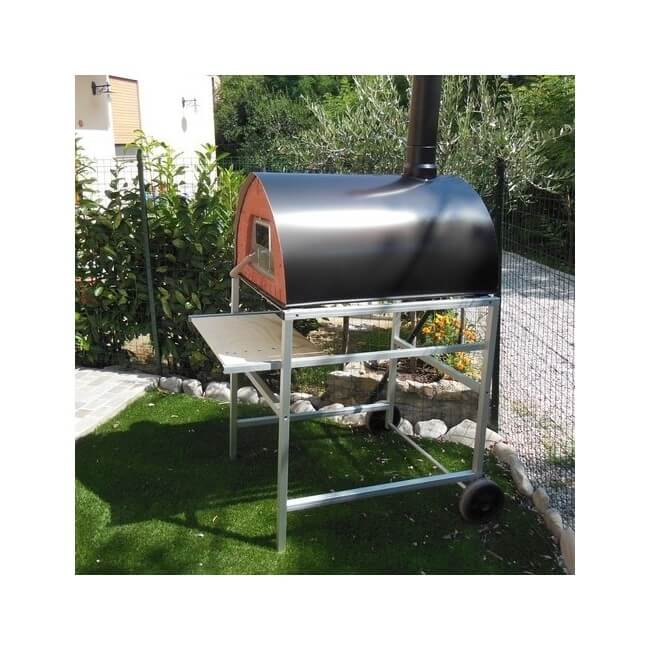A Pizza Party Passione Dual Fuel Pizza Oven sits on a black stand with a stainless steel shelf and wheels by Pizza Party, placed on artificial grass near garden pizzeria furniture, a stone path, and lush greenery.