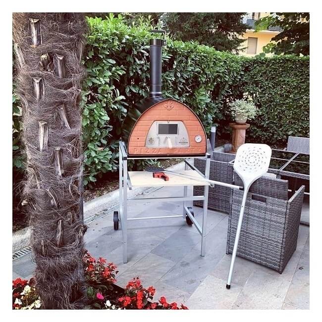 An outdoor pizza oven on a metal stand with a brick-look front panel, set on a patio next to a palm tree trunk, wicker furniture, and green hedges.