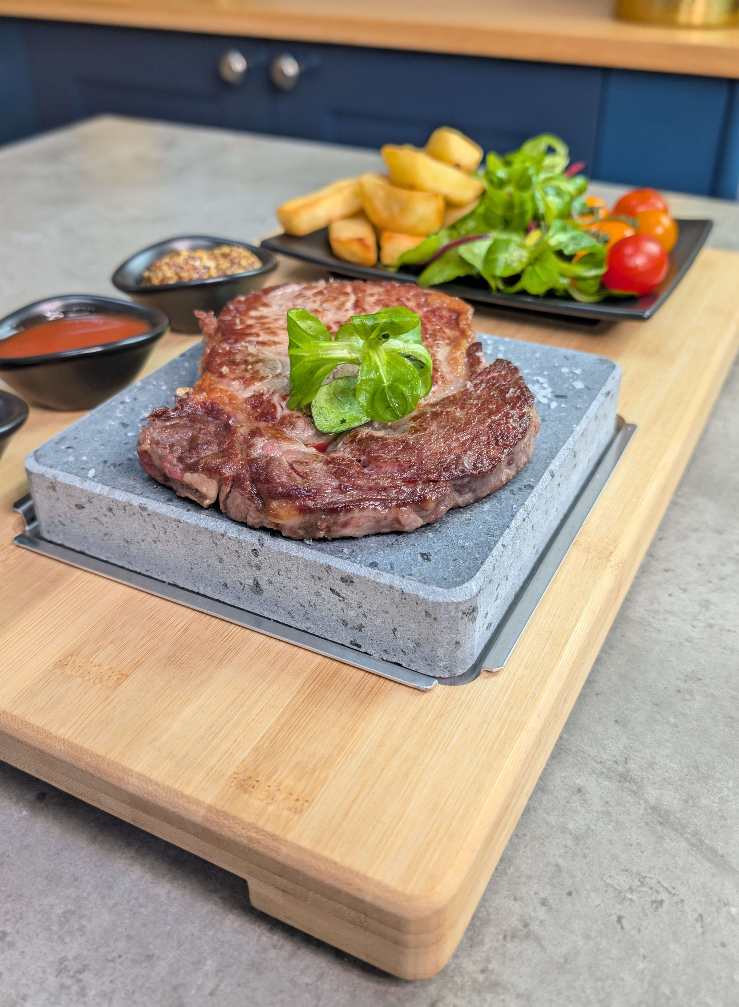 Angle shot of a Black Rock Grill HO-09PMB steak stone set featuring a ribeye steak chips and salad on a matt black side plate in a modern blue kitchen