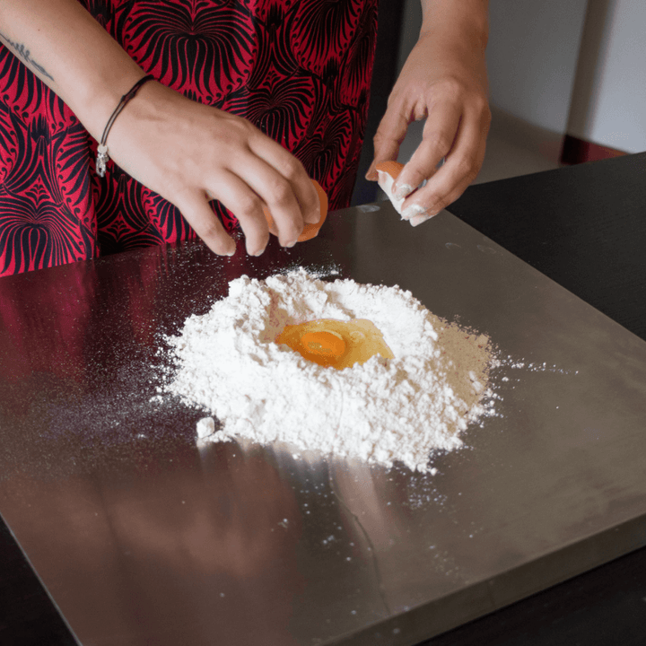 Wearing a red and black patterned apron, the person cracks an egg into a well of flour on their Effeuno Stainless Steel Pizza Prep Board—Professional Food-Grade AISI 304, 50cm, a perfect accessory for pizza-making.