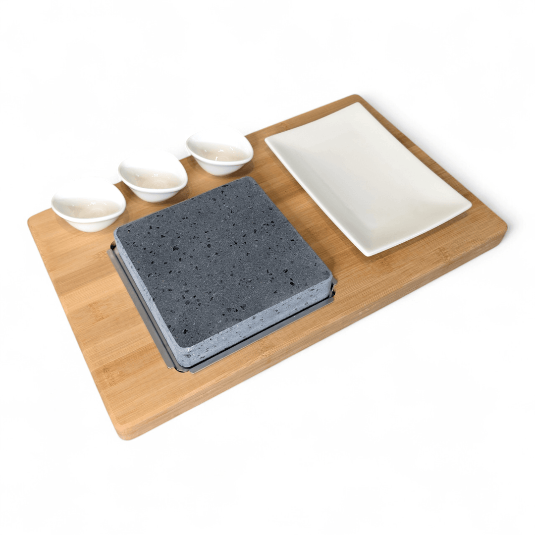 A bamboo serving board viewed sideways set on a white background, featuring a square lava cooking stone, a rectangular white side plate and three small white dipping pots.