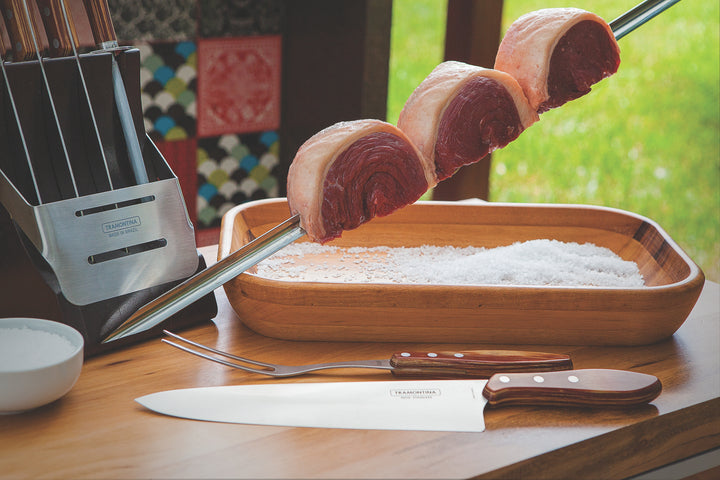 The Tramontina Churrasco BBQ Skewer 750mm, made of stainless steel, holds sliced beef above a wooden tray filled with coarse salt. Next to it is a Brazilian hardwood knife block with two knives. A small bowl of salt sits beside this setup against a blurred green outdoor background.