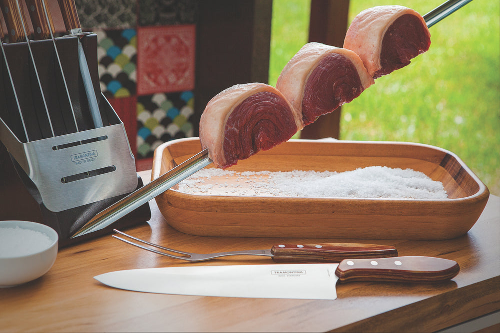 A Tramontina Churrasco BBQ Wide Skewer 750mm holds three pieces of raw meat above a wooden tray filled with coarse salt. A knife with a Brazilian hardwood handle is in a block on the table, with greenery in the background.
