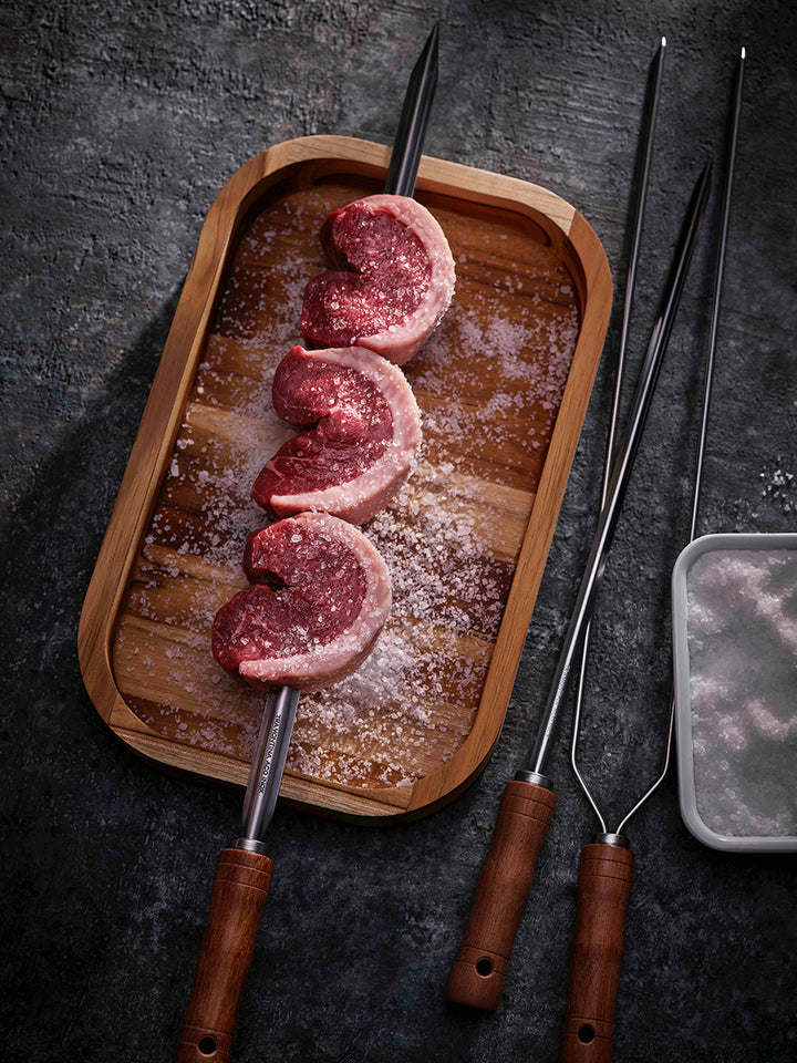 Tramontina Churrasco BBQ Skewers (750mm) with beef cuts rest on a wooden tray sprinkled with coarse salt; two additional Tramontina skewers and a salt container lie nearby on a textured dark surface.