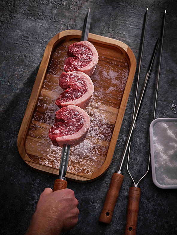 Three uncooked beef picanha steaks are skewered on the Tramontina Churrasco 850mm Wide BBQ Skewer, placed on a wooden tray with coarse salt. A hand holds one skewer while two additional skewers, featuring Brazilian hardwood handles, lie nearby next to a container of salt.