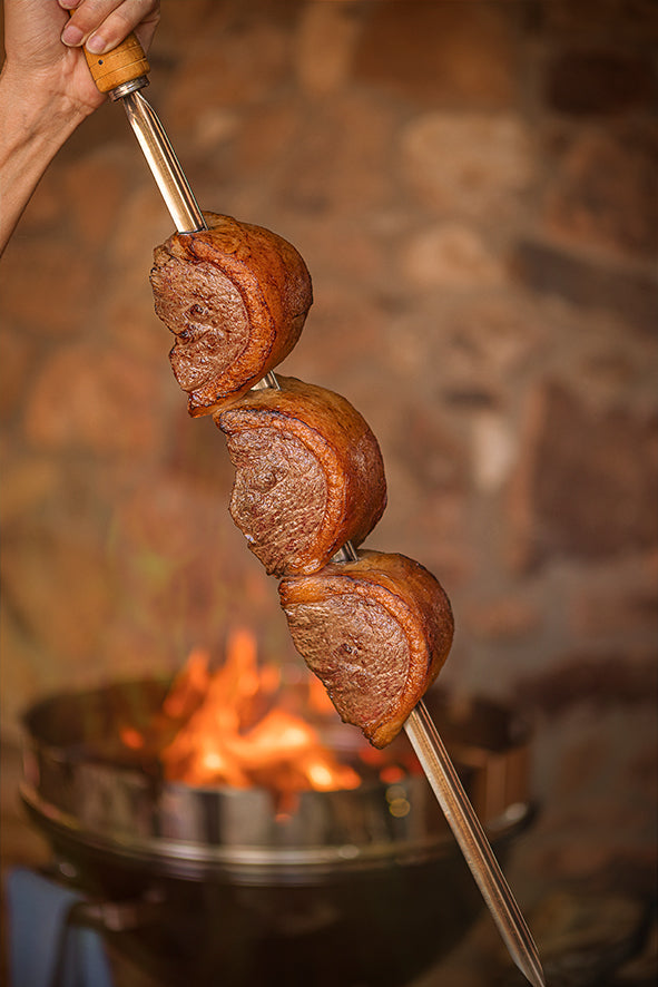 Three slices of grilled steak are skewered on a Tramontina Churrasco BBQ Skewer 750mm, held over a barbecue grill with visible flames in the background, evoking a cozy outdoor cooking setting.