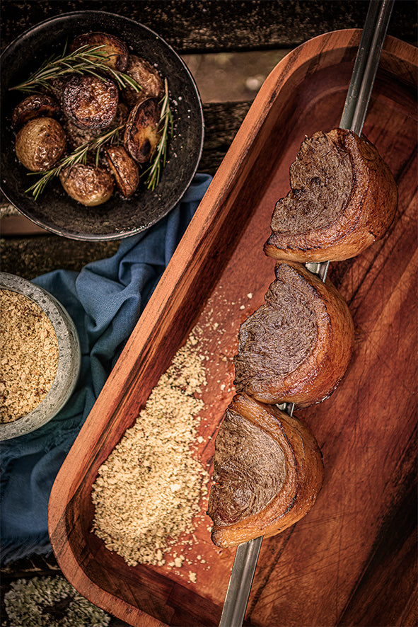 Close-up of a Tramontina Churrasco 850mm Wide BBQ Skewer holding three grilled meat pieces on a wooden tray with scattered breadcrumbs. A bowl of rosemary roasted potatoes and a small bowl of breadcrumbs are in the background, enhancing the rustic charm.