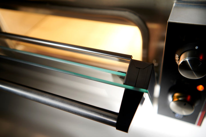 Close-up of the open Black Rock Grill Effeuno P134H 509 Manual Pizza Oven, featuring a glass panel, metal handle, manual Neapolitan-style controls and indicator lights on the right. The interior light is on, illuminating the oven.