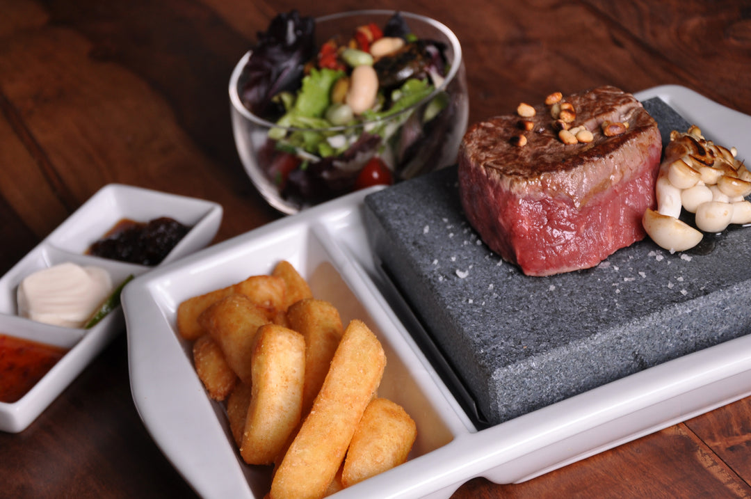 Enjoy a gourmet meal with a rare steak sizzling on a cooking stone, served with roasted garlic, thick-cut fries, three dipping sauces, and a mixed salad of greens and beans—all presented on Black Rock Grills Premium White Porcelain Steak Stone Platters- GP-10.