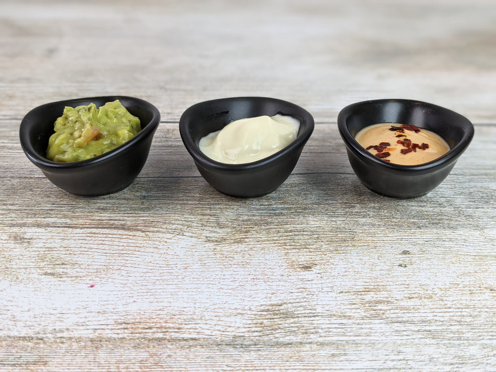 Three small ramekins from the Black Rock Grill 12 Matt Black Porcelain Ramekin Dipping Pots set rest on a wooden surface, holding guacamole, creamy white sour cream, and a tan sauce garnished with red pepper flakes.
