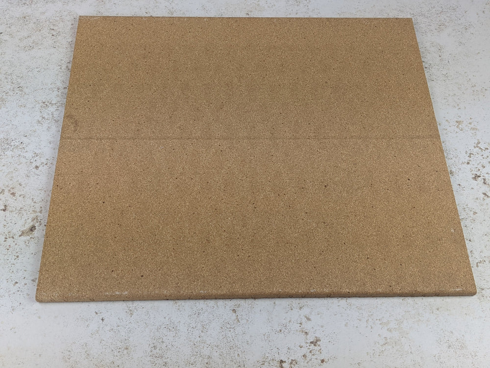 A square corkboard lies on a light, speckled surface similar to the texture of a Black Rock Grill Refractory Stone for Pizza Oven. The board is plain with a subtle seam across the middle.