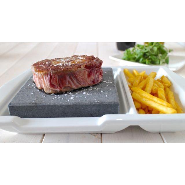 A seared steak on a Black Rock Grill Steak Stone Plate, sprinkled with salt, is served on a porcelain plate with fries. A side of greens is blurred in the background on a wooden table for healthy dining, showcasing the Black Rock Grill dining experience.